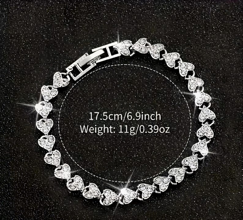 May include: A silver bracelet featuring heart-shaped links, each encrusted with small, sparkling crystals. The bracelet is 6.9 inches long and weighs 0.39 ounces. The clasp is visible, and the bracelet is displayed on a dark surface.