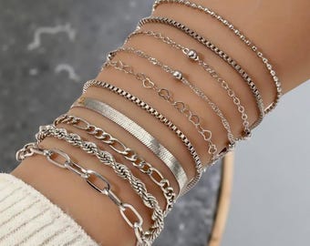 May include: A collection of silver bracelets in various styles, including chain, rope, and heart-shaped designs. The bracelets are layered on a wrist, showcasing a mix of textures and patterns. The jewelry is displayed against a neutral background.