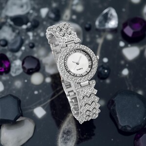 May include: A silver quartz watch with a round face and a band adorned with sparkling crystals. The watch face is white with black Roman numerals and hands. The watch is set against a dark, reflective surface with decorative stones.