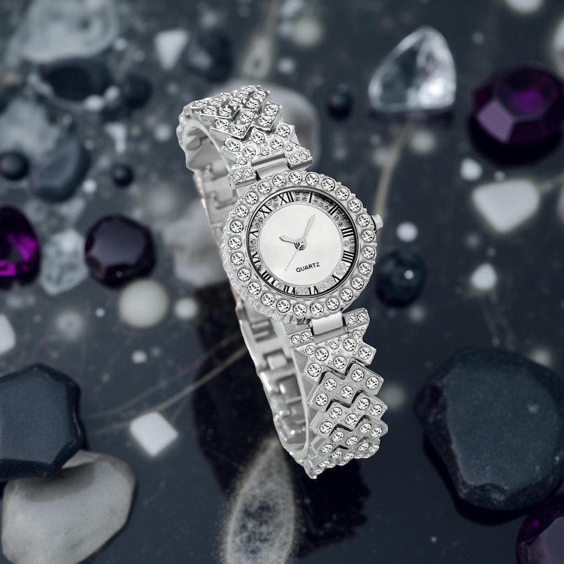May include: A silver quartz watch with a round face and a band adorned with sparkling crystals. The watch face is white with black Roman numerals and hands. The watch is set against a dark, reflective surface with decorative stones.