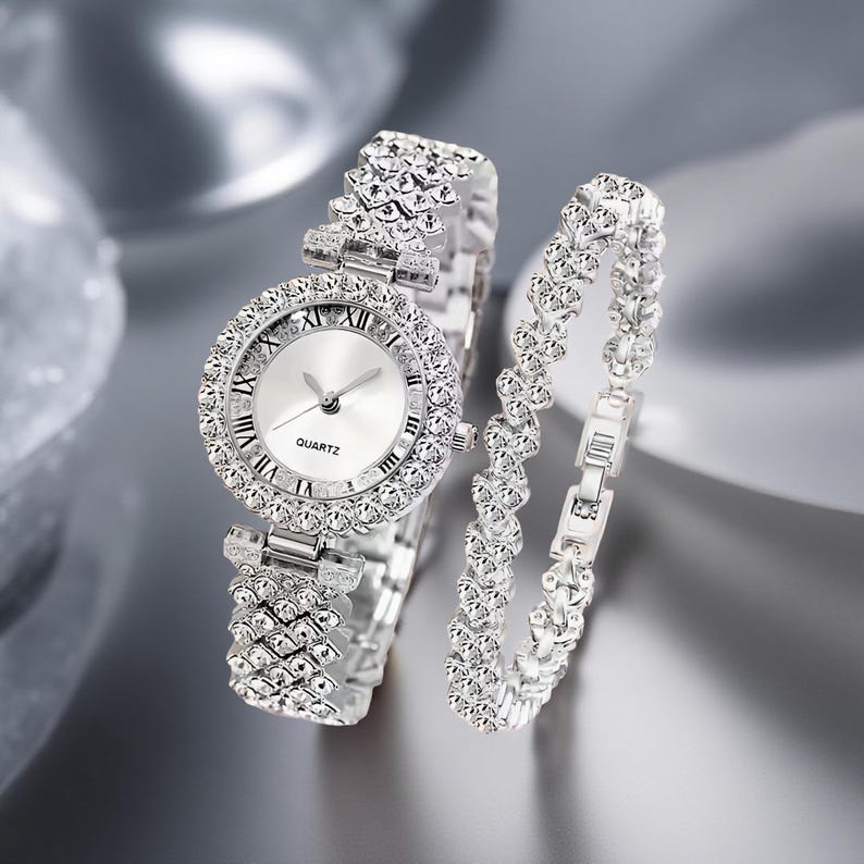 May include: A silver-toned quartz watch and matching bracelet set. The watch features a round face with Roman numeral hour markers and a bezel encrusted with clear crystals. The bracelet is also adorned with crystals.