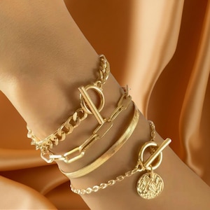 Może przedstawiać: Zestaw bransoletek w kolorze złota z różnymi stylami łańcuszków. Bransoletki obejmują gruby łańcuch typu curb, łańcuch z prostokątnymi ogniwami, łańcuch w jodełkę i łańcuch z koralików z zawieszką w kształcie monety. Każda bransoletka ma unikalne zapięcie.