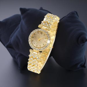 May include: A gold-toned wristwatch with a round face and a band adorned with sparkling crystals. The watch face features gold-colored hands and hour markers. The watch is displayed on a dark blue surface.