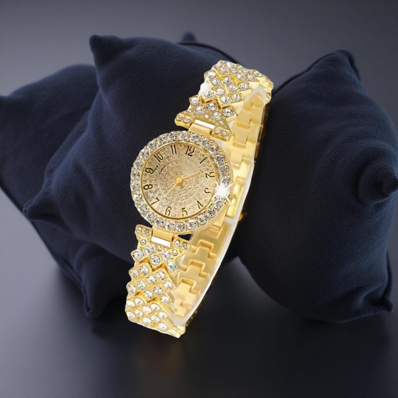 May include: A gold-toned wristwatch with a round face and a band adorned with sparkling crystals. The watch face features gold-colored hands and hour markers. The watch is displayed on a dark blue surface.