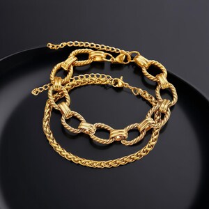 May include: Two gold-colored bracelets with different chain designs. The top bracelet features large, oval links, while the bottom bracelet has a smaller, braided chain. Both bracelets have a shiny finish and are displayed on a black surface.