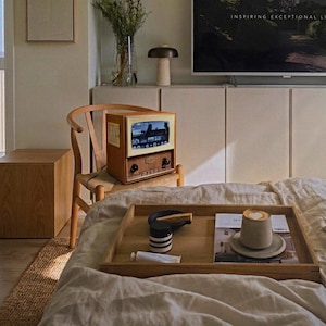 Può includere: Un vassoio di legno con una tazza di caffè, una rivista e un contenitore a strisce bianche e nere è appoggiato su un letto. Un televisore in stile vintage è su una sedia di legno. Il televisore ha un piccolo schermo.