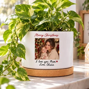 May include: A white ceramic planter on a round cork base containing a variegated green and yellow pothos plant. The planter features a printed photograph of two people, the text Merry Christmas, and the handwritten text I love you Mom Love, Olivia.