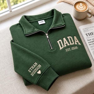 May include: A dark green quarter-zip sweatshirt featuring a fold-over collar and a silver zip. The chest displays embroidered beige text reading DADA EST. 2026. The sleeve features embroidered beige text reading ETHAN HUDSON with a small heart.
