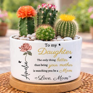 May include: A white ceramic planter with a variety of cacti, including one with an orange flower. The planter has a floral design and text that reads "To my Daughter". It sits on a round wooden base and a woven mat.