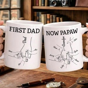 May include: Two white ceramic mugs with black text and illustrations. One mug reads "FIRST DAD" and the other reads "NOW PAPAW". Both mugs feature a design of hands holding smaller hands, with names below. The mugs are held by two people.