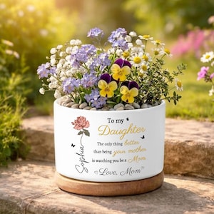 May include: A white ceramic planter with a wooden base, filled with colorful flowers. The planter has text that reads "To my Daughter" and "Love, Mom". A rose and the name "Sophia" are also featured.