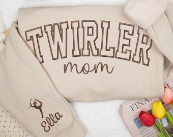 May include: A beige sweatshirt with the words "TWIRLER mom" in brown lettering. The left sleeve features a silhouette of a baton twirler and the name "Ella". The sweatshirt is folded on a white surface.