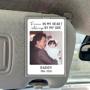 May include: A rectangular car visor clip featuring a photograph of an adult and a child. The text reads Forever IN MY HEART Always BY MY SIDE, DADDY, and 1964-2026. The clip is made of clear acrylic and is attached to a gray vehicle sun visor.