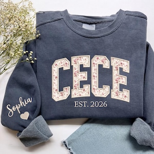 May include: A dark blue sweatshirt with the name "CECE" in a floral design. The text "EST. 2026" is below the name. The left sleeve has the name "Sophia" embroidered with a heart.