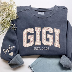 May include: A charcoal gray crewneck sweatshirt featuring the word GIGI in large block letters with a pink and white floral pattern. Below the text is EST. 2026. The left sleeve displays the name Sophia in white script above a small heart icon.