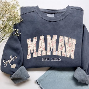 May include: A dark blue sweatshirt with the word "MAMAW" in a floral design across the chest. The text "EST. 2026" is below the word. The sleeve has the name "Sophia" embroidered with a heart.