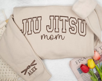 以下が含まれることがあります： ベージュ色のスウェットシャツ。茶色で「JIU JITSU mom」の文字が刺繍されています。左袖には茶色のベルトのデザインがあり、その下に「ALEX」という名前が刺繍されています。スウェットシャツは白い表面に折りたたまれています。