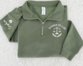 May include: Olive green quarter-zip sweatshirt. The left sleeve has the name "Sophia" embroidered in white, along with a small heart. The right chest features the text "WOMEN IN LAW" with a scale of justice emblem.
