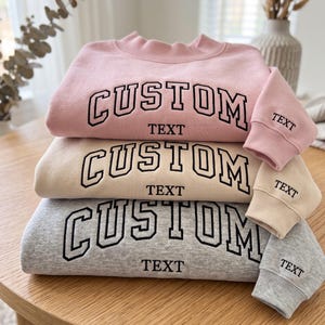 May include: A stack of three crewneck sweatshirts in light pink, beige, and heather gray. Each sweatshirt features the word CUSTOM in large black collegiate-style lettering across the chest and the word TEXT embroidered on the sleeve cuff.
