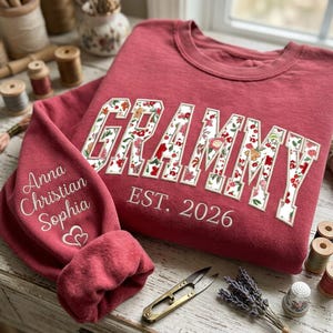 Peut inclure: Un sweat-shirt à col rond rose poudré avec le mot GRAMMY en grandes lettres majuscules à motif floral sur la poitrine. En dessous, le texte EST. 2026 est brodé. La manche affiche les prénoms Anna, Christian et Sophia avec deux cœurs.