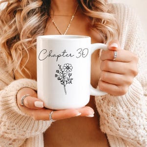 May include: White ceramic mug with the text "Chapter 30" in a handwritten font. A black floral illustration is below the text. The mug has a large handle and is held by a person.