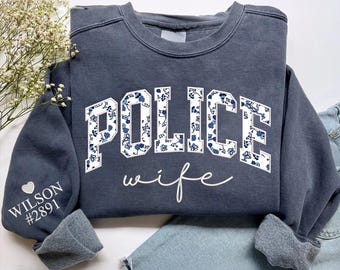 May include: A charcoal gray crewneck sweatshirt featuring the word POLICE in block letters with a blue and white floral pattern, and the word wife in white script below. The left sleeve displays the text WILSON #2891 with a small heart icon.