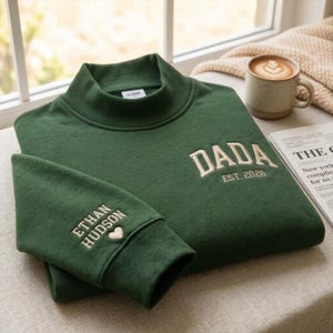 May include: A dark green mock neck sweatshirt featuring cream-coloured embroidery. The chest displays the text DADA EST. 2026, while the sleeve cuff is embroidered with the name ETHAN HUDSON and a small heart symbol. The garment has a ribbed collar.