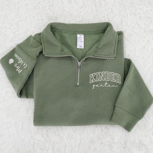 May include: Olive green quarter-zip sweatshirt with the words "Kindergarten" in white script across the chest. The left sleeve has the text "Mrs. Wilson" in white script. The sweatshirt has a collared neckline and a silver zipper.