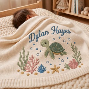 May include: A cream-colored knit baby blanket featuring an embroidered design of a green sea turtle, pink and green coral, a yellow starfish, and blue bubbles. The name Dylan Hayes is embroidered in blue script text above the sea turtle.