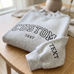 May include: A light gray crewneck sweatshirt featuring the word CUSTOM in large black embroidered block letters across the chest, with the word TEXT embroidered below it and again on the sleeve in a smaller black font with ribbed cuffs.
