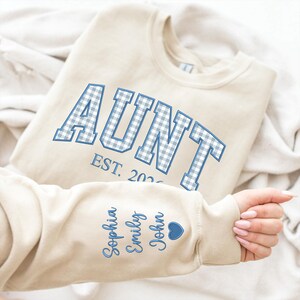 May include: A beige crewneck sweatshirt featuring the word AUNT in blue and white gingham block letters with the text EST. 2026 underneath. The sleeve is embroidered with the names Sophia, Emily, and John in blue script next to a blue heart.