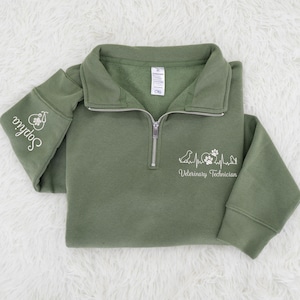 May include: Olive green quarter-zip sweatshirt with white embroidered text and graphics. The left sleeve has the name "Sophia" and a paw print design. The front features a heartbeat line, paw prints, and the words "Veterinary Technician".