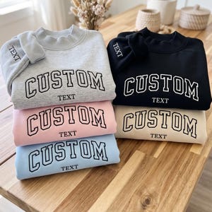 May include: A stack of five crewneck sweatshirts in light gray, black, light pink, light blue, and cream. Each sweatshirt features the word CUSTOM in a large arched font and the word TEXT below it, with TEXT also embroidered on the sleeve.