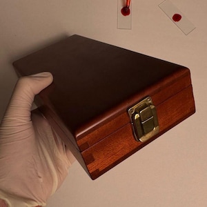 Peut inclure: Une boîte en bois foncé avec un fermoir en laiton est tenue par une main gantée. Plusieurs lames de microscope en verre avec des gouttelettes de liquide rouge sont dispersées sur une surface blanche. Une main gantée tient une pipette avec du liquide rouge.
