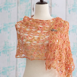 May include: A peach and gold crocheted shawl draped over a mannequin. The open weave design features a delicate pattern, secured with a decorative brooch. The shawl's colors include shades of peach, gold, and a hint of red.