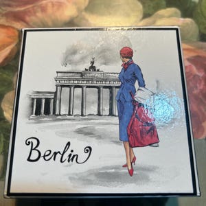 May include: Square decorative box with a black border featuring a watercolor illustration of the Brandenburg Gate in Berlin. A woman in a blue suit and red accessories stands in front of the gate. The word "Berlin" is written in cursive.