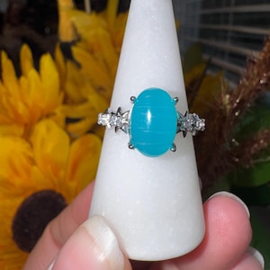K&ouml;nnte beinhalten: Ein silberfarbener Ring mit einem gro&szlig;en, ovalen, leuchtend t&uuml;rkisfarbenen Cabochon-Stein mit horizontaler B&auml;nderung. Die Ringschiene ist auf beiden Seiten der Fassung mit kleinen, klaren, funkelnden Steinen in sternf&ouml;rmigem Metall verziert.