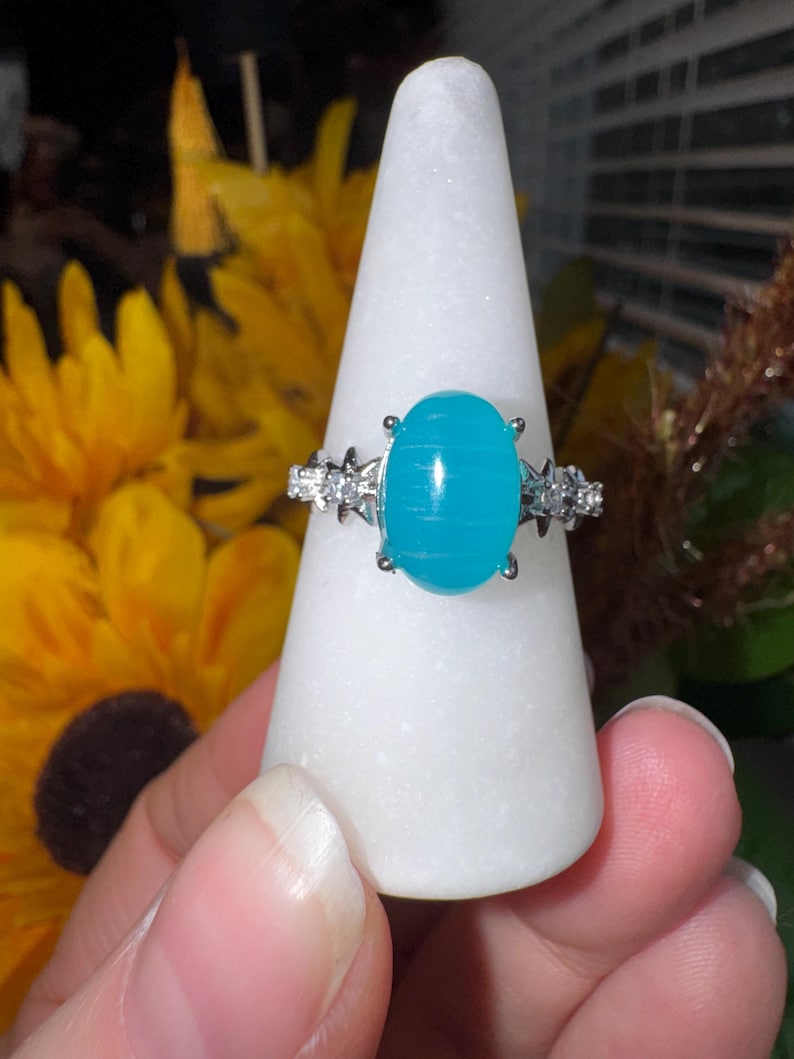 K&ouml;nnte beinhalten: Ein silberfarbener Ring mit einem gro&szlig;en, ovalen, leuchtend t&uuml;rkisfarbenen Cabochon-Stein mit horizontaler B&auml;nderung. Die Ringschiene ist auf beiden Seiten der Fassung mit kleinen, klaren, funkelnden Steinen in sternf&ouml;rmigem Metall verziert.