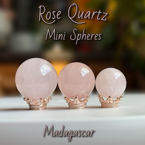 May include: Three rose quartz mini spheres on decorative gold-colored stands. The spheres vary in shades of pink and are labeled "Rose Quartz Mini Spheres" and "Madagascar".