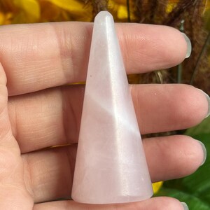 K&ouml;nnte beinhalten: Ein glatter, blassrosa Rosenquarzkristall in konischer Form. Der Stein hat ein durchscheinendes Aussehen mit subtilen internen Variationen und einer polierten Oberfl&auml;che, die sich zu einer abgerundeten Spitze und einer flachen Basis verj&uuml;ngt.