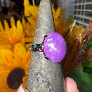 K&ouml;nnte beinhalten: Ein Ring mit einem gro&szlig;en, ovalen violetten Stein mit wei&szlig;en Einschl&uuml;ssen, gefasst in ein dunkles Metallband. Das Band ist mit kleinen, eingebetteten gr&uuml;nen und roten Akzentsteinen an der Seite verziert und weist eine detaillierte, texturierte Metalloberfl&auml;che auf.