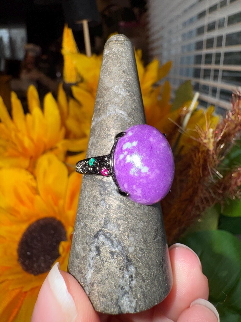 K&ouml;nnte beinhalten: Ein Ring mit einem gro&szlig;en, ovalen violetten Stein mit wei&szlig;en Einschl&uuml;ssen, gefasst in ein dunkles Metallband. Das Band ist mit kleinen, eingebetteten gr&uuml;nen und roten Akzentsteinen an der Seite verziert und weist eine detaillierte, texturierte Metalloberfl&auml;che auf.