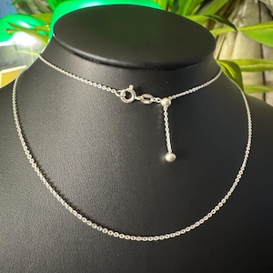 May include: A delicate silver necklace with a fine chain and adjustable length. The necklace is displayed on a black mannequin, showcasing its simple elegance. The chain features a small, round charm at the end of the adjustable portion.