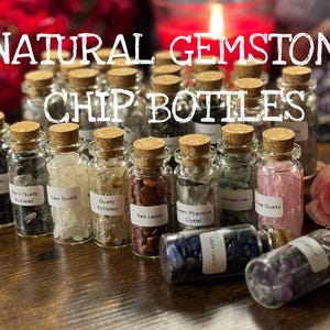 May include: A collection of small glass bottles filled with various natural gemstone chips, each labeled with the gemstone type. The bottles have cork stoppers and are arranged on a wooden surface. The text "NATURAL GEMSTONE CHIP BOTTLES" is at the top.
