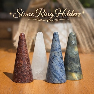May include: Four cone-shaped stone ring holders arranged in a row. The first is reddish-brown, the second is translucent white, the third is blue with dark speckles, and the fourth is grey with iridescent flashes. Text reads: Stone Ring Holders.