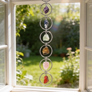 May include: A vertical hanging ornament featuring seven silver-toned metal rings linked together in a chain. Each ring contains a single raw, unpolished gemstone in a different color, including clear, purple, blue, green, brown, pink, and red stones.
