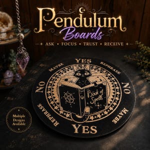May include: A round black pendulum board featuring gold-colored engravings of a cat reading a book titled Book of Spells, with text reading YES, NO, MAYBE, and REPHRASE. A crystal pendulum hangs on a silver chain to the left of the board.