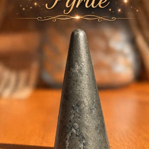 K&ouml;nnte beinhalten: Ein kegelf&ouml;rmiges Objekt aus metallisch grauem Pyrit mit einer glatten, polierten Oberfl&auml;che und sichtbaren nat&uuml;rlichen Mineraleinschl&uuml;ssen. Das Wort Pyrite ist in einer kursiven, goldfarbenen Schrift &uuml;ber dem Stein gedruckt, erg&auml;nzt durch kleine dekorative Funken.