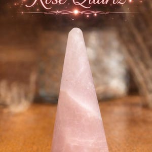 K&ouml;nnte beinhalten: Ein blassrosa Rosenquarzkristall, der in eine glatte, konische Form geschliffen ist. Der Stein weist nat&uuml;rliche interne Maserungen und ein halbtransparentes Aussehen auf. &Uuml;ber dem Kristall steht der Text Rose Quartz in wei&szlig;er Schrift.