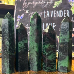 May include: Five polished ruby zoisite crystal towers. The crystals are a mix of dark green and black with red inclusions. The crystals are standing on a wooden surface. A chalkboard with text is in the background.